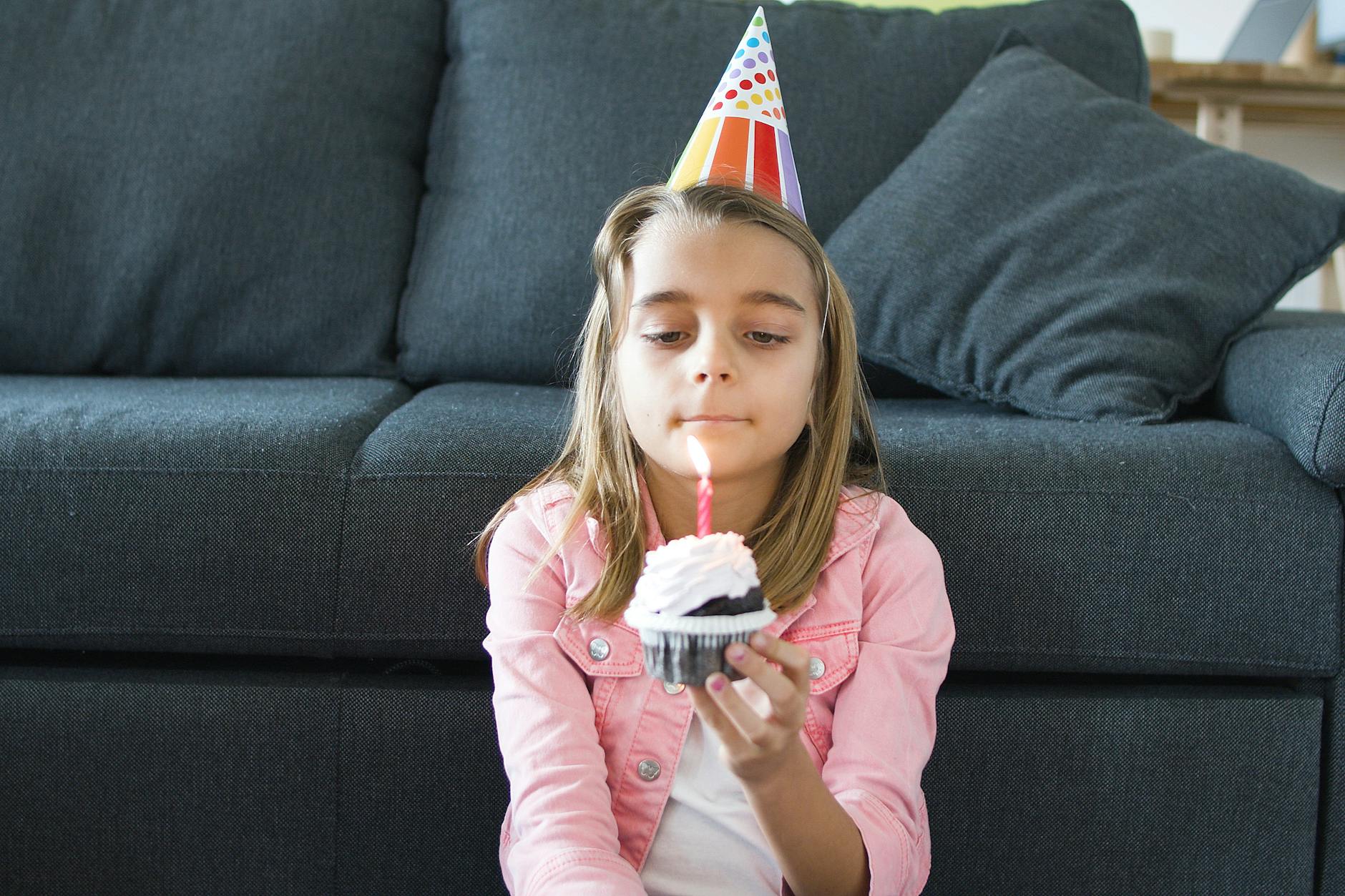 Dziewczynka w czapce urodzinowej trzyma babeczkę z zapaloną świeczką